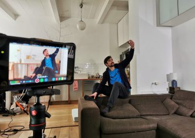 a man sits on the back of the couch and in the foreground we can see him in a camera monitor