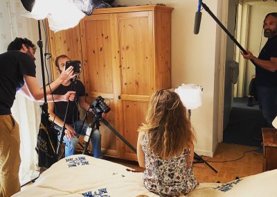 Camera man and assistant ready a camera as a sound man holds a boom mic and the actress sits on the bed waiting