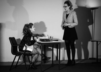 One actress seated at a desk on stage as the another actress stands on the other side of the desk